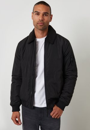 Schwarze Bomberjacke mit Reißverschluss, Sherpa-Kragen, gerippten Bündchen und Seitentaschen; getragen über einem weißen Shirt und grauen Jeans.