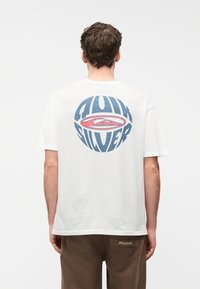 Άνδρας γυρισμένος με την πλάτη φοράει λευκό μπλουζάκι με μπλε και κόκκινο λογότυπο Quiksilver και καφέ παντελόνι με τη λέξη «PEGADOR» στην πίσω τσέπη.