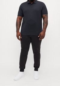 Man som bär svart polo-shirt, svarta joggerbyxor och vita sneakers, stående mot en enkel vit bakgrund.