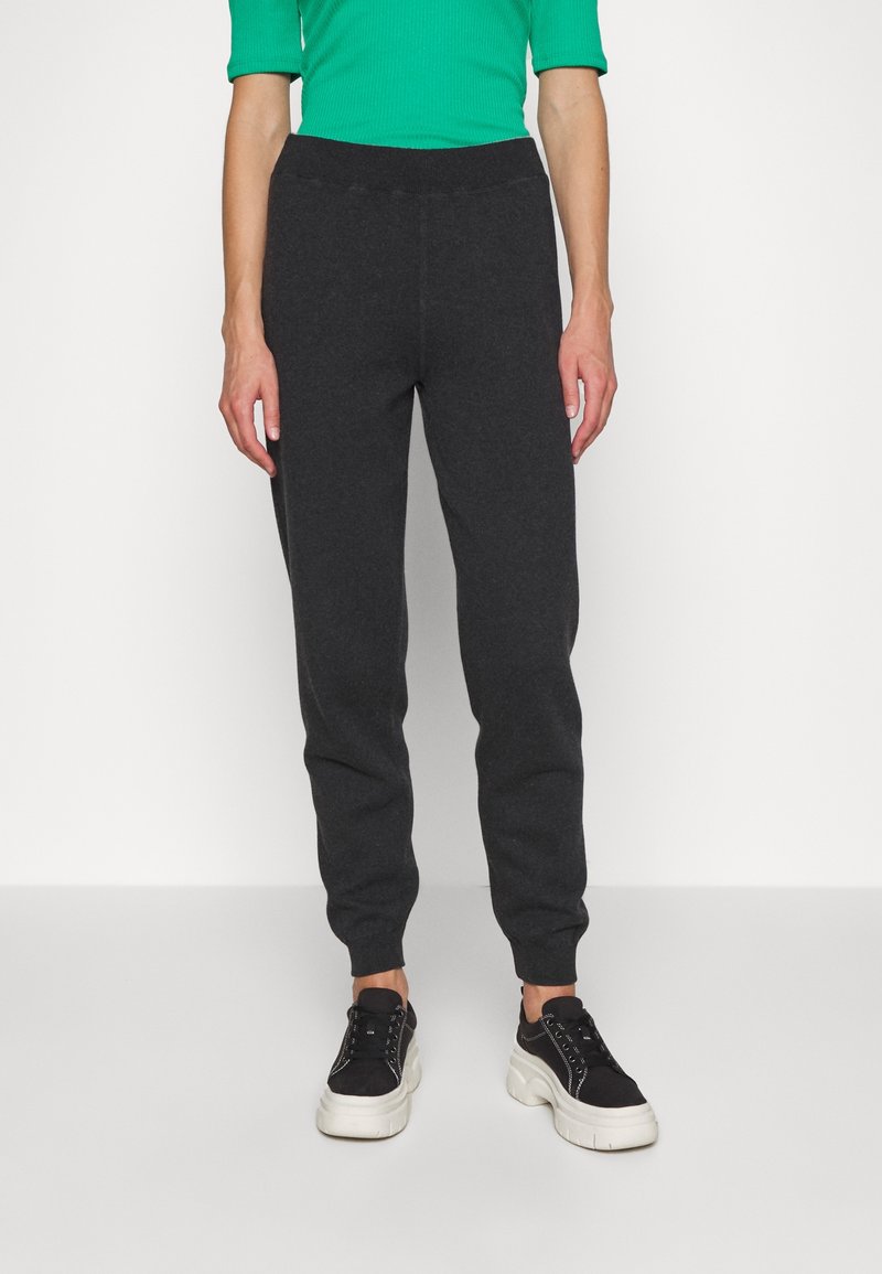 Personne portant un pantalon de jogging gris foncé, des baskets noires avec des semelles blanches épaisses, et un haut à manches longues vert fluo, se tenant devant un fond blanc.