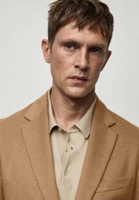 Svijetlo smeđi vuneni sako preko bež polo majice. Sako ima klasične reveri i jedan džep na prsima. Glatka tekstura, čiste linije.