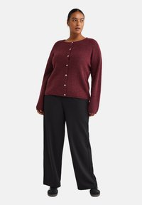 Maglione a maglia bordeaux con maniche lunghe e bottoni frontali, abbinato a pantaloni neri larghi e scarpe nere. Tessuto semplice e texturizzato.