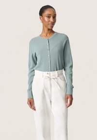 Cardigan a coste verde chiaro con scollatura rotonda e chiusura con bottoni, indossato sopra pantaloni bianchi a gamba larga con texture.