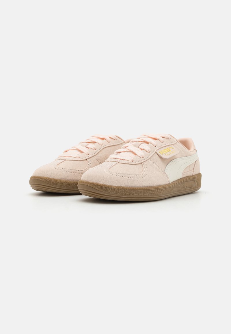 Baskets en daim rose clair avec une rayure latérale blanche, lacets plats et semelle en caoutchouc marron texturé. Présente une étiquette logo sur le côté.