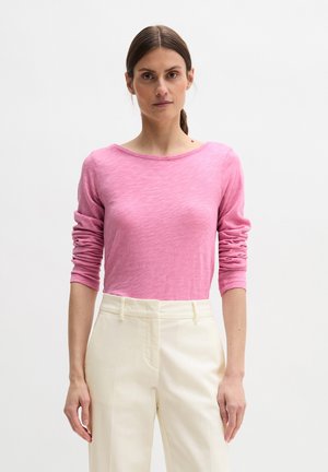 Kobieta o brązowych włosach spiętych do tyłu, ubrana w różową bluzkę z długim rękawem i białe spodnie z wysokim stanem, stojąca na jednolitym białym tle.