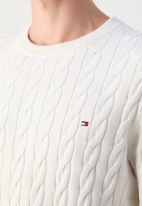 Gros plan d'un pull blanc en maille torsadée avec col rond et petit logo rouge, blanc et bleu sur la poitrine, porté par une personne.
