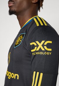 Black sports jersey with yellow accents, featuring a round collar and three stripes on the shoulder. Logo on chest and sleeve. Textured fabric.