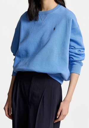 Mujer que lleva una sudadera azul clara de manga larga con un pequeño logo oscuro en el pecho izquierdo, combinada con pantalones negros de talle alto.