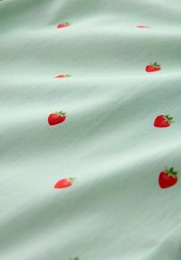 Tejido verde claro con pequeños estampados de fresas rojas dispersos y pliegues sutiles que crean suaves sombras.