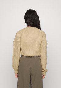 Femme aux longs cheveux foncés portant un pull court côtelé beige et un pantalon taille haute vert olive, debout devant un fond uni.