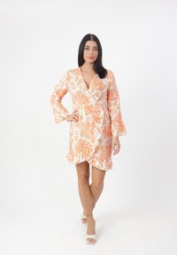 Bloemenwikkeljurk in wit en oranje, met lange klokmouwen, een gerimpelde zoom en een V-hals. Gemaakt van lichtgewicht stof.