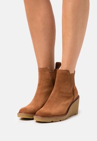 Brown suede wedge ankle boots worn on bare legs with textured rubber soles and pull tabs at the back.