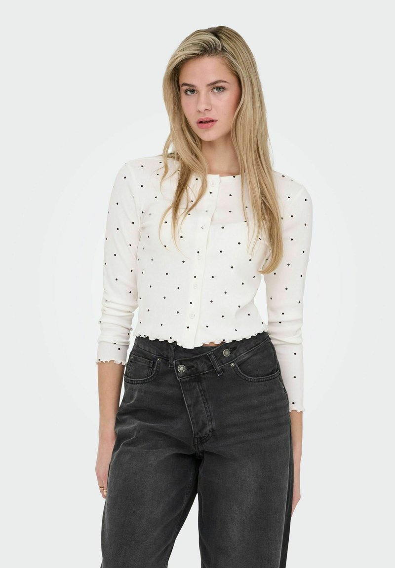 Cardigan blanc à côtes avec des pois noirs, encolure ronde, ourlet festonné et manches longues, associé à un jean noir taille haute.