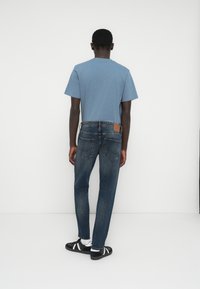 Μπλε κοντομάνικη μπλούζα, τζιν παντελόνι με καφέ δερμάτινο ταμπελάκι και μαύρα σανδάλια με λευκές ρίγες. Απλό λευκό φόντο.