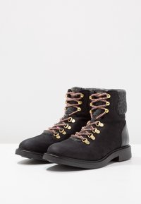 Schwarze Wildleder-Wanderschuhe mit einem grauen Filzkragen, goldenen Ösen und pinken Schnürsenkeln. Flache Sohle und abgerunde Zehenform für Stabilität und Komfort.