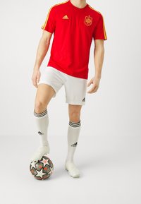 Rode Adidas voetbalshirt met gouden accenten, witte shorts en zwartgestreepte witte sokken. Speler staat op een zwart-wit gestippelde voetbal.