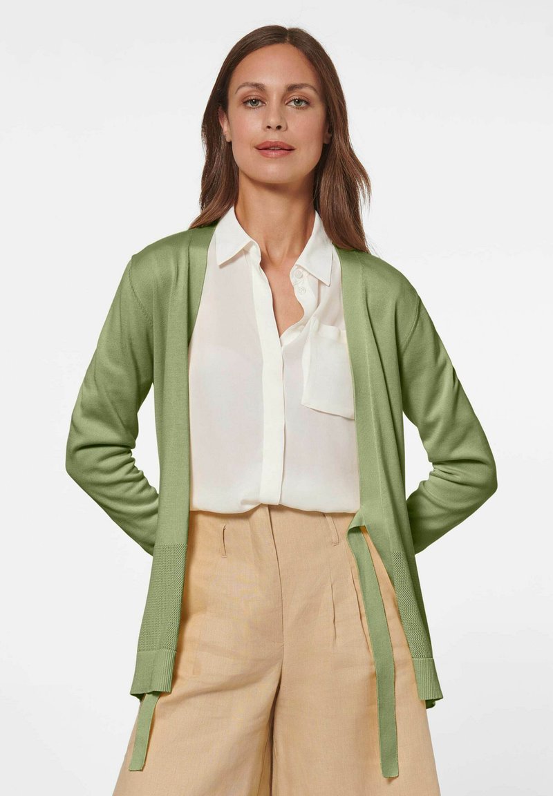 Lichtgroene cardigan van zachte stof, met een open voorkant, lange mouwen en zijknopen, gecombineerd met een witte blouse en beige broek.