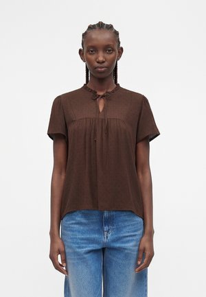 Vrouw met gevlochten haar die een bruine blouse met korte mouwen en een striksluiting bij de hals en een blauwe spijkerbroek draagt, staande tegen een effen witte achtergrond.