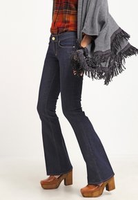 Dunkelblaue Schlagjeans mit niedriger Taille und kontrastierenden Nähten, kombiniert mit braunen Plateauschuhen. Ein gefranster grauer Poncho rundet den Look ab.