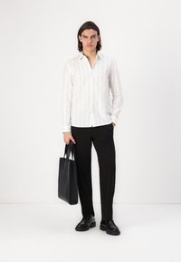 Witte blouse met lange mouwen en verticale strepen, zwarte broek en zwarte leren schoenen. Houdt een zwarte tote bag vast. Minimalistisch ontwerp.