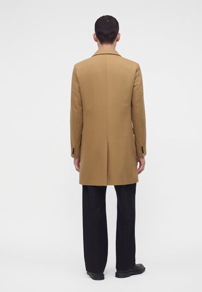Homme debout de dos, portant un long manteau couleur chameau, un pantalon noir et des chaussures noires, sur un fond blanc uni.