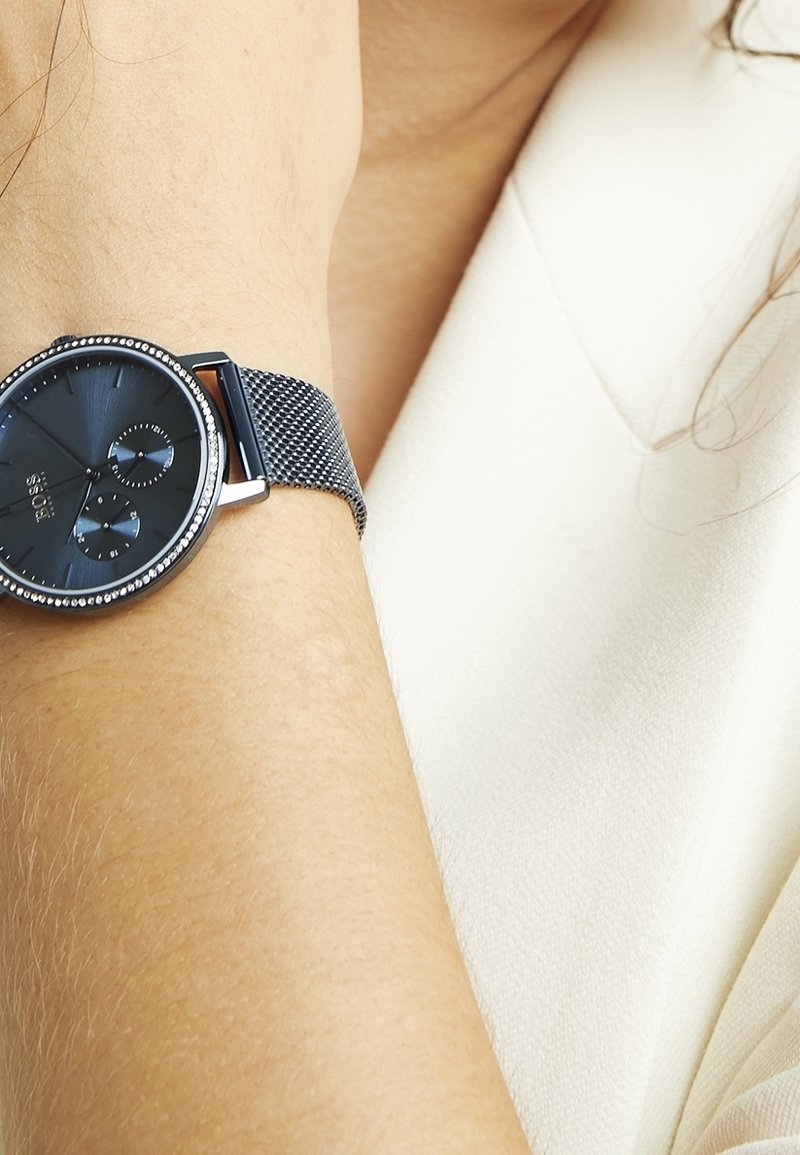 Poignet portant une montre ronde noire avec un bracelet en maille métallique et deux sous-cadrans, sur peau claire et fond en tissu blanc.
