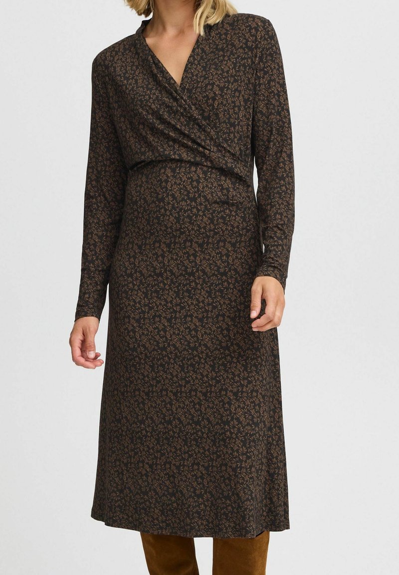 Femme portant une robe midi manches longues marron foncé avec un petit motif de feuilles et un col croisé, associée à des bottes marron.