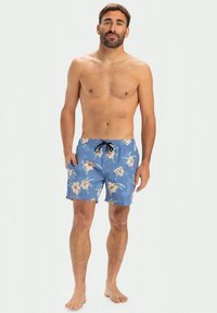 Muž bez trička stojí na boso, oblečený v modrých kvetinových plavkách, s rukami v vreckách na bielom pozadí.