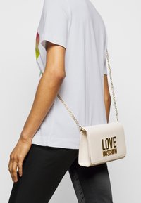 Béžová umělá kožená kabelka přes rameno se zlatým nápisem "LOVE MOSCHINO", má řetízek a obdélníkový tvar.