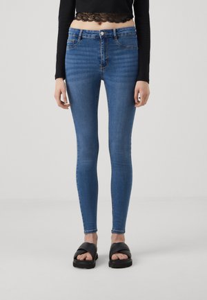 Slim-fit, korkeavyötäröiset siniset farkut, joissa on klassinen viisi-tasku design ja lievää kulumista tekstuurin vuoksi. Yhdistetty mustiin sandaaleihin.