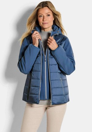 Donna che chiude una giacca trapuntata blu sopra un maglione grigio, indossa pantaloni beige, in piedi contro uno sfondo chiaro e uniforme.