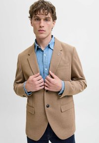 Beige strukturierter Blazer mit zwei Fronttaschen und einem einzelnen Knopfverschluss, darüber ein hellblaues Denim-Hemd. Das Model posiert mit den Händen auf dem Blazer.