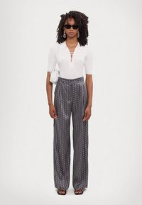 Kvinne med krøllete hår som har på seg svarte solbriller, hvit kortermet bluse, grå bukser med polka-dot mønster og vid ben, svarte sandaler og en hvit skulderveske.