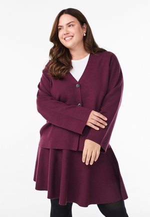 Smilende kvinde med langt brunt hår iført en blommefarvet cardigan med knapper over et matchende nederdel og hvid top, stående mod en hvid baggrund.