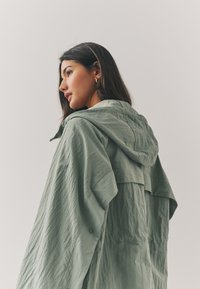 Light green hooded jacket made of textured fabric, featuring a loose fit, a layered back design, and a snap button detail on the sleeve.