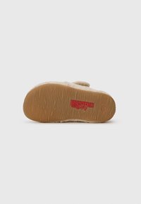 Semelle de chausson en feutre beige avec caoutchouc texturé marron. Présente un logo rouge et un symbole circulaire, des bords cousus et un détail de bande surélevée.
