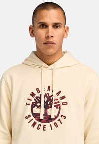 Felpa beige con tasca frontale a canguro, cappuccio con cordino e un logo circolare che presenta un design ad albero e la scritta "TIMBERLAND SINCE 1973" in bordeaux.