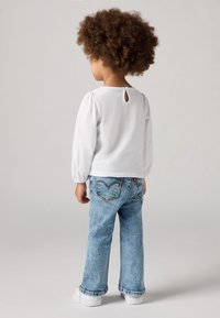 Camicia bianca a maniche lunghe con dettaglio di bottoni sul retro, abbinata a jeans in denim chiaro a zampa d'elefante con cuciture a contrasto e sneakers bianche.