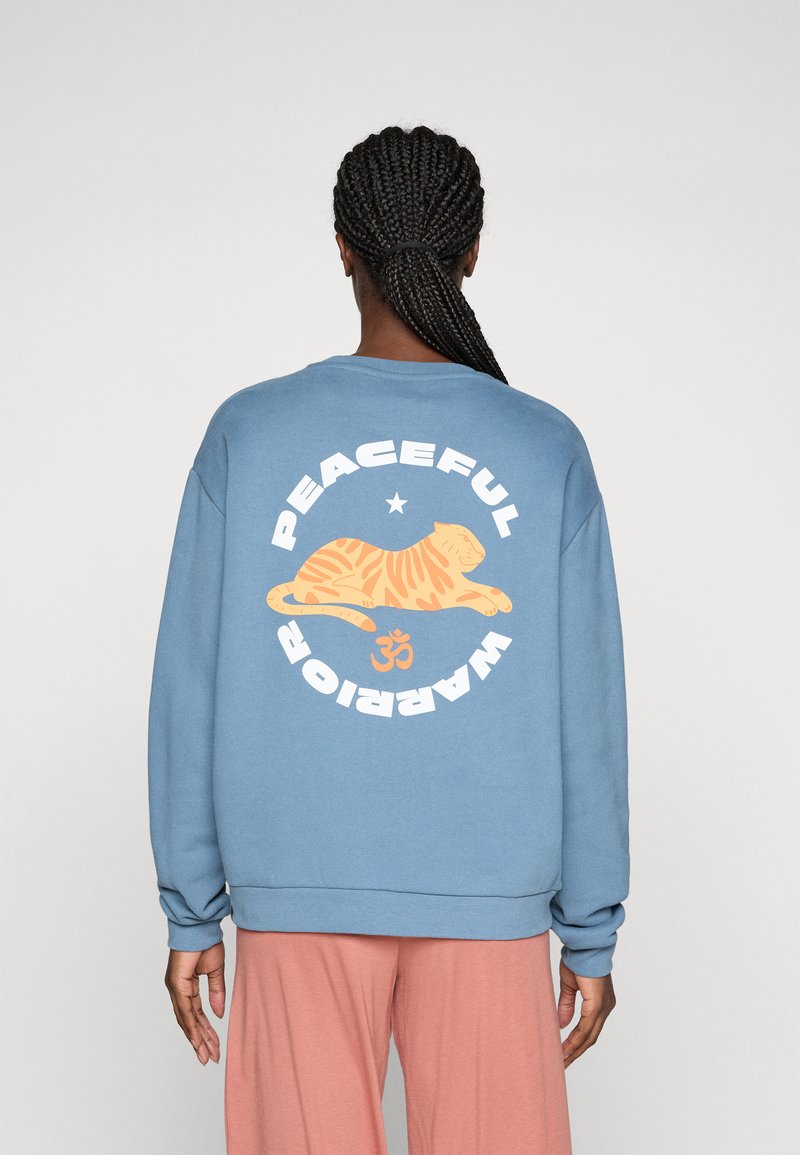Persona con capelli intrecciati che indossa una felpa blu con un tigre arancione e la scritta "Guerriero Pacifico" sulla schiena, abbinata a pantaloni rosa.