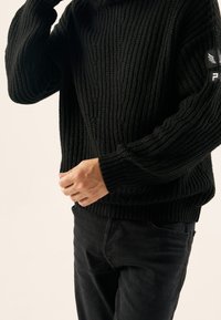 Svart ribbad stickad tröja med långa ärmar, hög krage och texturerade detaljer. Bärs med mörka jeans, som har ett märke på vänster arm.