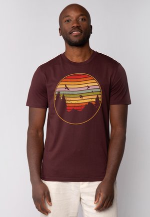 Bordeauxrotes Baumwoll-T-Shirt mit einem kreisförmigen Grafikdesign, das horizontale Streifen in Orange, Gelb, Grün und Rot zeigt, sowie Bäume und Vögel darstellt.