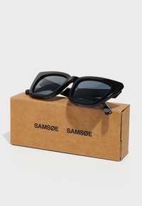 Ochelari de soare cu ramă neagră în formă de pătrat și lentile întunecate, așezați pe o cutie de carton bej etichetată "SAMSOE." Designul are forme groase și unghiulare.