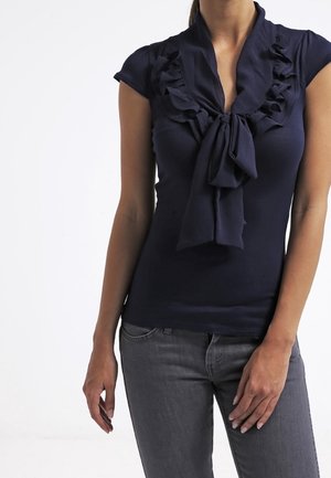 Marineblaue Bluse mit kurzen Ärmeln, die Rüschen am Kragen und eine gebundene Schleife am Hals hat. Figurbetonter Schnitt kombiniert mit grauen Jeans.