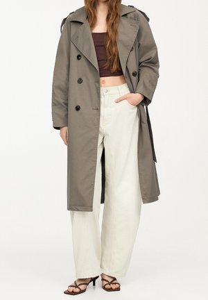 Vrouw die een lange grijze trenchcoat draagt, een donkerbruin cropped topje, witte broek met wijde pijpen en bruine sandalen met hak en bandjes.