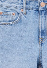 Jean en denim bleu clair en gros plan montrant la poche avant, la passants de ceinture, les rivets en cuivre et le bouton en métal avec des détails de couture visibles.