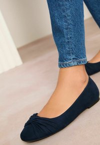 Ballerines en daim bleu marine avec un design noué au niveau des orteils. Forme de bout arrondi et semelle en caoutchouc plate, associées à un jean en denim bleu clair.