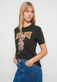 Camiseta de algodão cinzenta escura com um gráfico de um urso vestido de rosa, com o texto "New York" em fonte estilizada laranja. Mangas curtas.