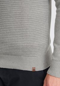 Gray knitted sweater featuring a textured pattern, ribbed hem and cuffs, and a small brown leather tag near the bottom seam.