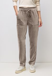 Frau trägt taupefarbene Cord-Hose mit Kordelzug, aufgesetzten Fronttaschen und weiße Sneaker, steht vor einem schlichten Hintergrund.