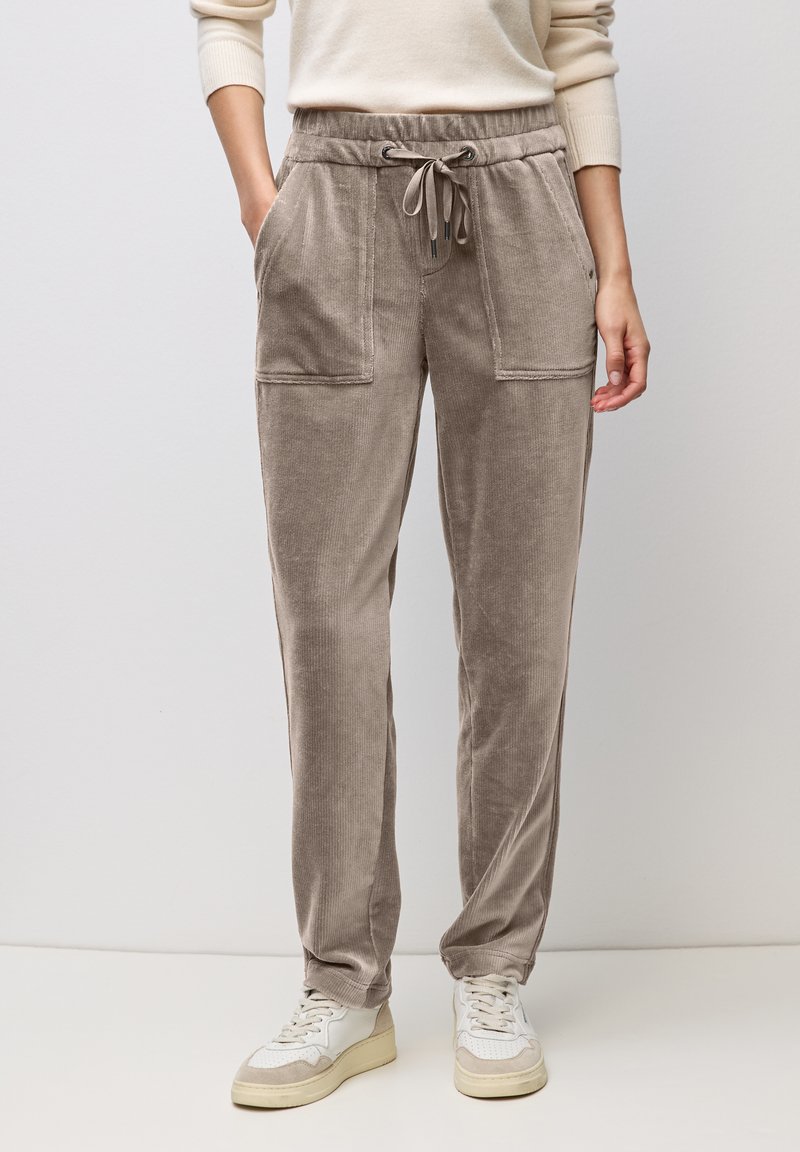 Frau trägt taupefarbene Cord-Hose mit Kordelzug, aufgesetzten Fronttaschen und weiße Sneaker, steht vor einem schlichten Hintergrund.